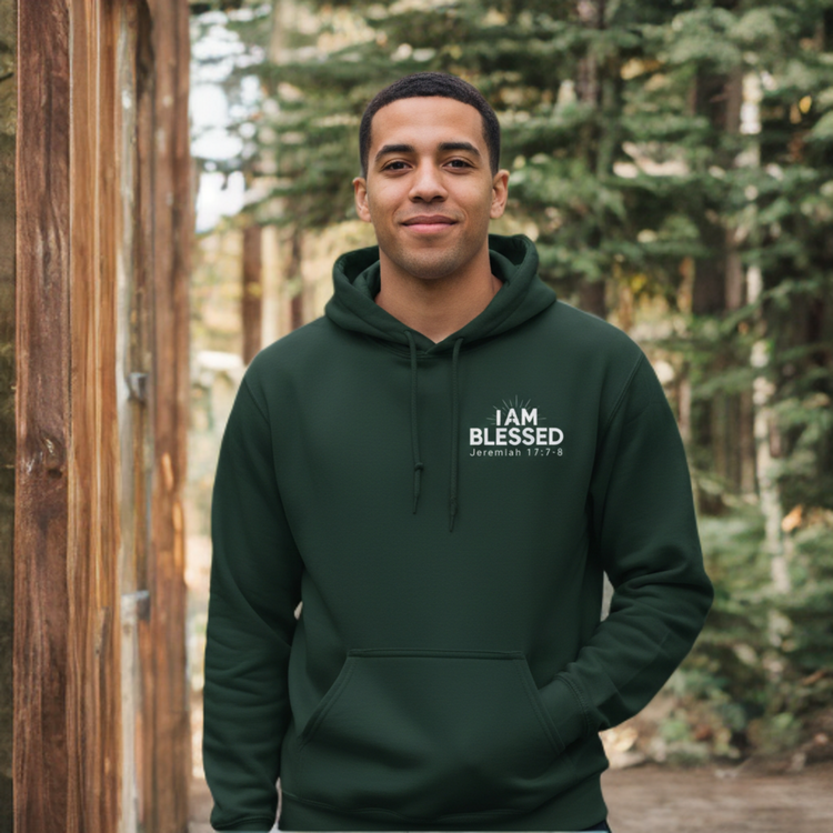 ​A smiling man standing in a pine forest wearing a dark forest green hoodie. The hoodie features a white graphic on the chest that reads "I AM BLESSED" with the scripture reference "JEREMIAH 17:7-8" underneath.