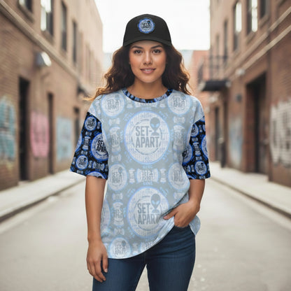 A woman in an alley wearing a black cap and a light blue t-shirt featuring a repeating "SET APART" logo with an alien icon.