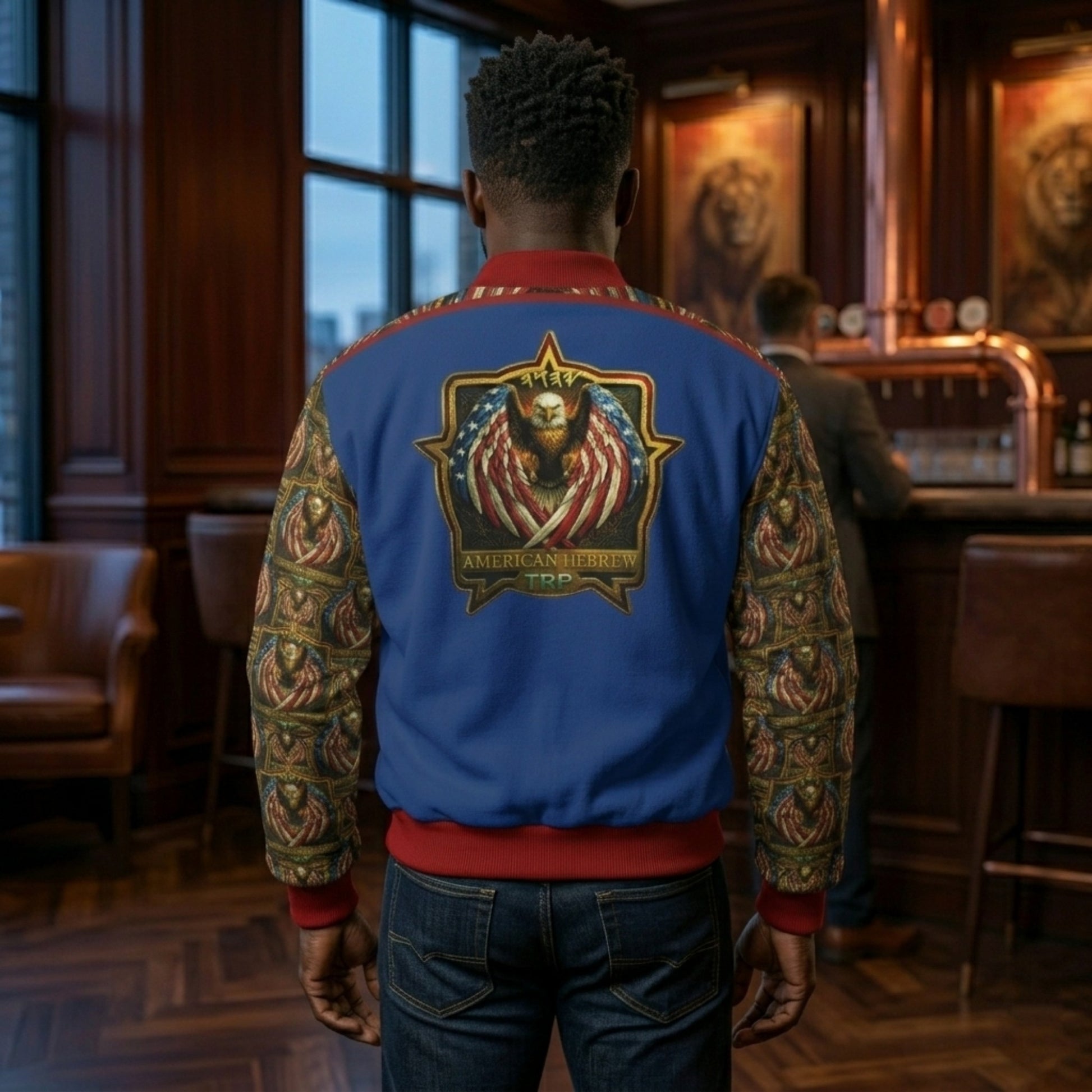 Rear view of a man wearing a blue bomber jacket with a large "American Hebrew" eagle emblem on the back and patterned sleeves, standing in a lounge.