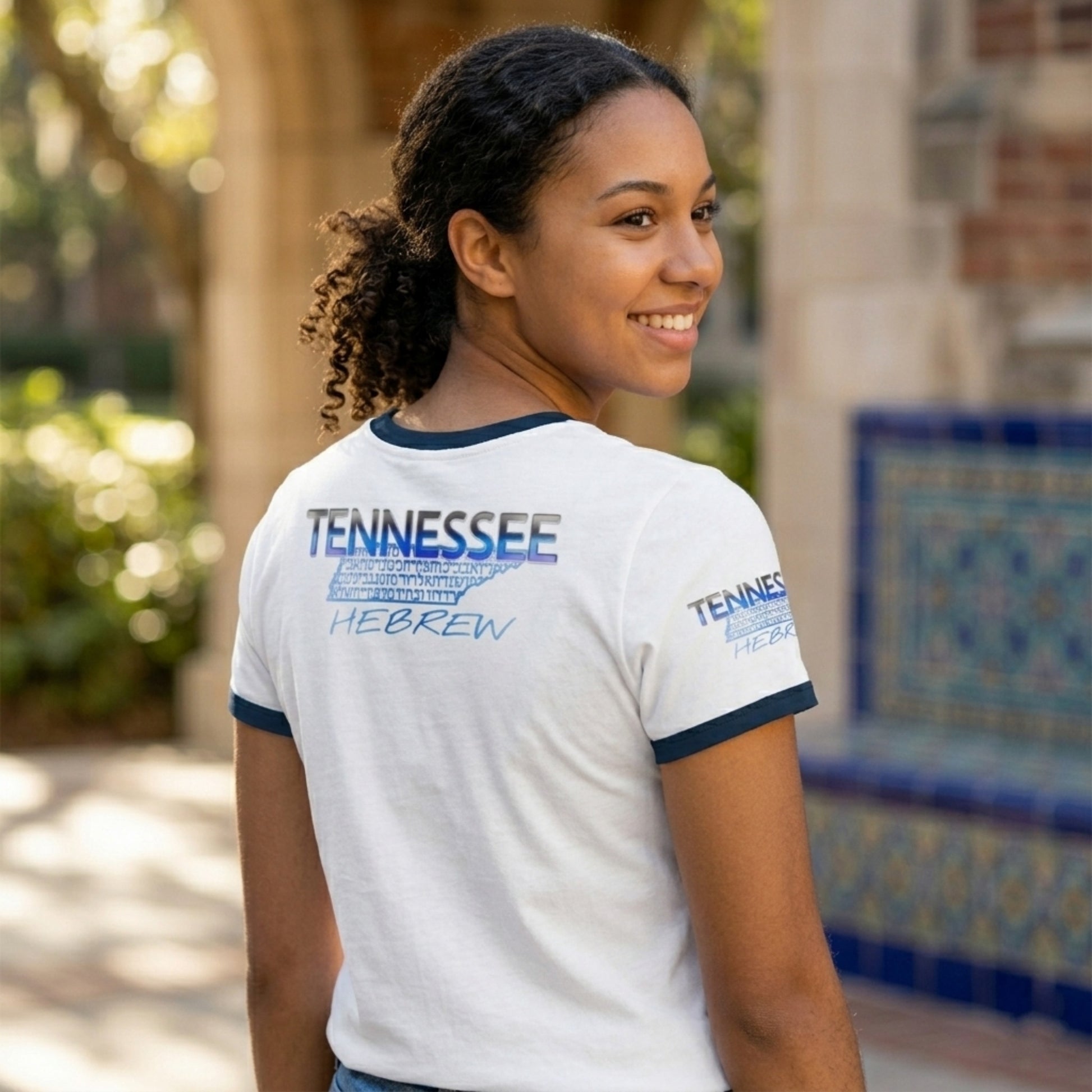 A woman looks over her shoulder, showcasing the back of a white ringer tee with a blue "TENNESSEE" logo, a state map in Hebrew script, and "HEBREW" below.