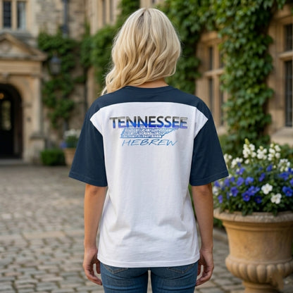 Rear view of a woman wearing a white and navy raglan shirt with a "TENNESSEE HEBREW" graphic on the upper back, standing in a stone courtyard.