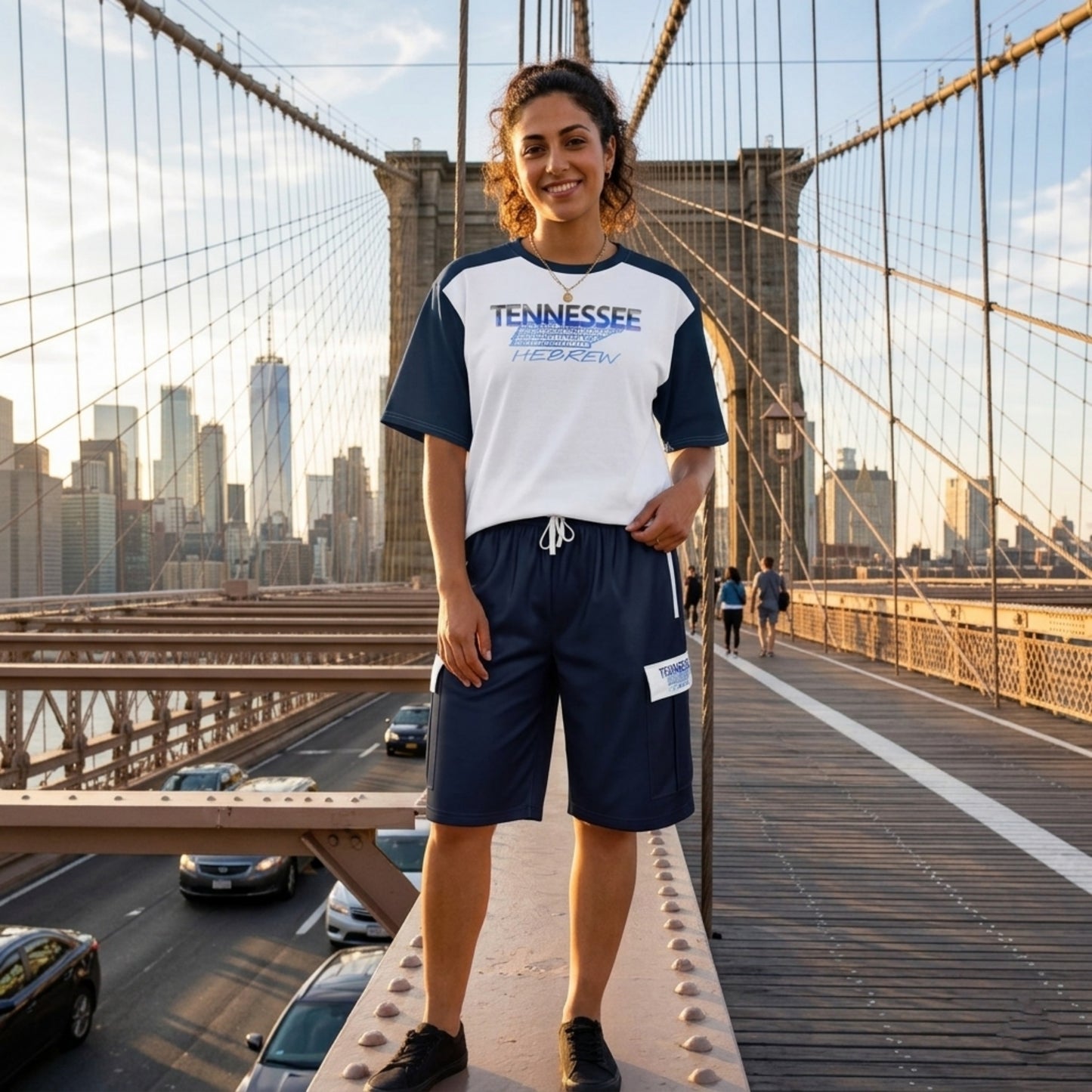 Woman smiling on the Brooklyn Bridge wearing a white and navy "Tennessee Hebrew" t-shirt with matching navy cargo shorts.