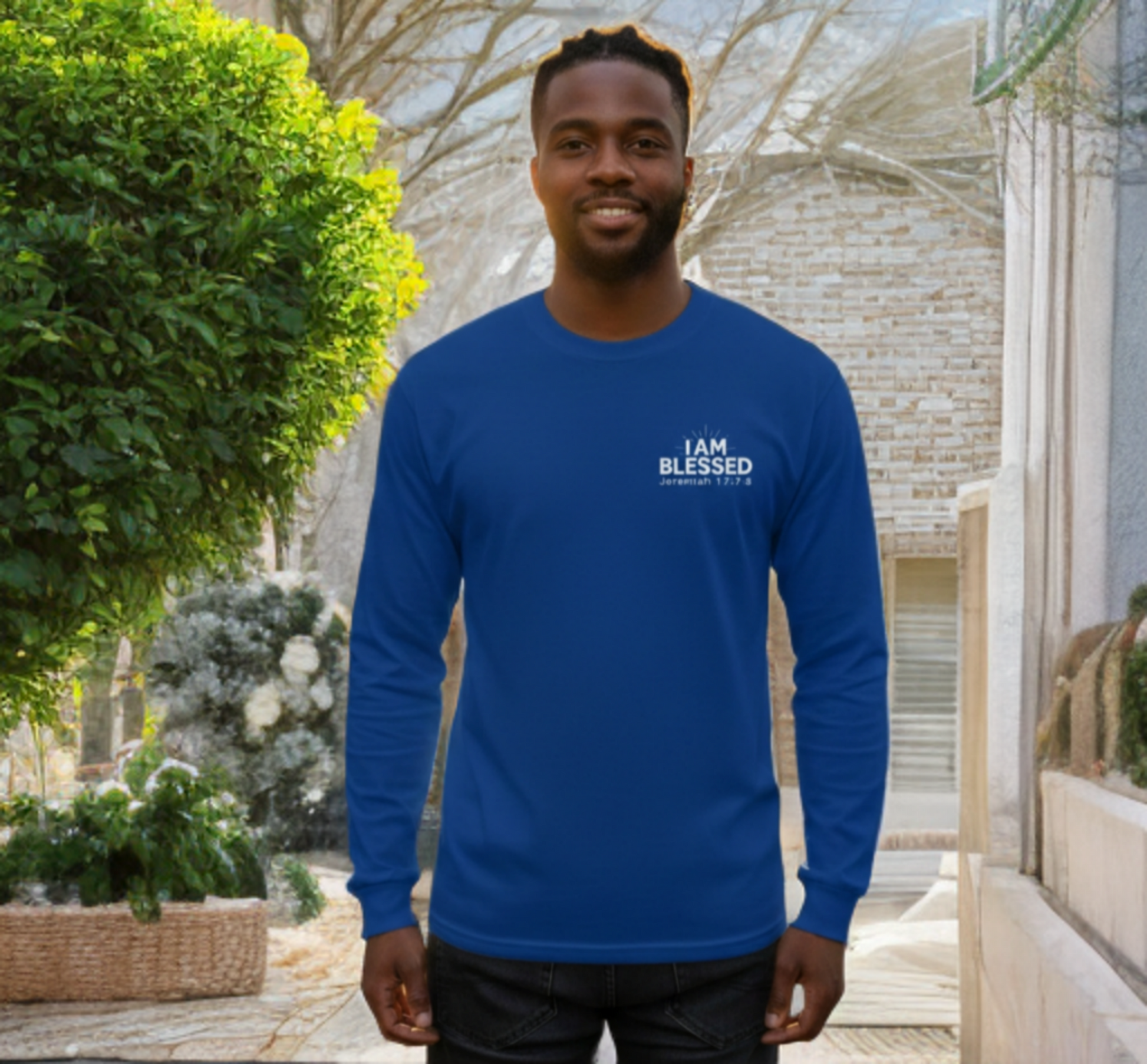 A medium shot of a smiling Black man standing outdoors, wearing a royal blue long-sleeved t-shirt. On the left chest of the shirt, there is a white graphic that reads "I AM BLESSED" above the scripture reference "Jeremiah 17:7-8." He is also wearing dark jeans.