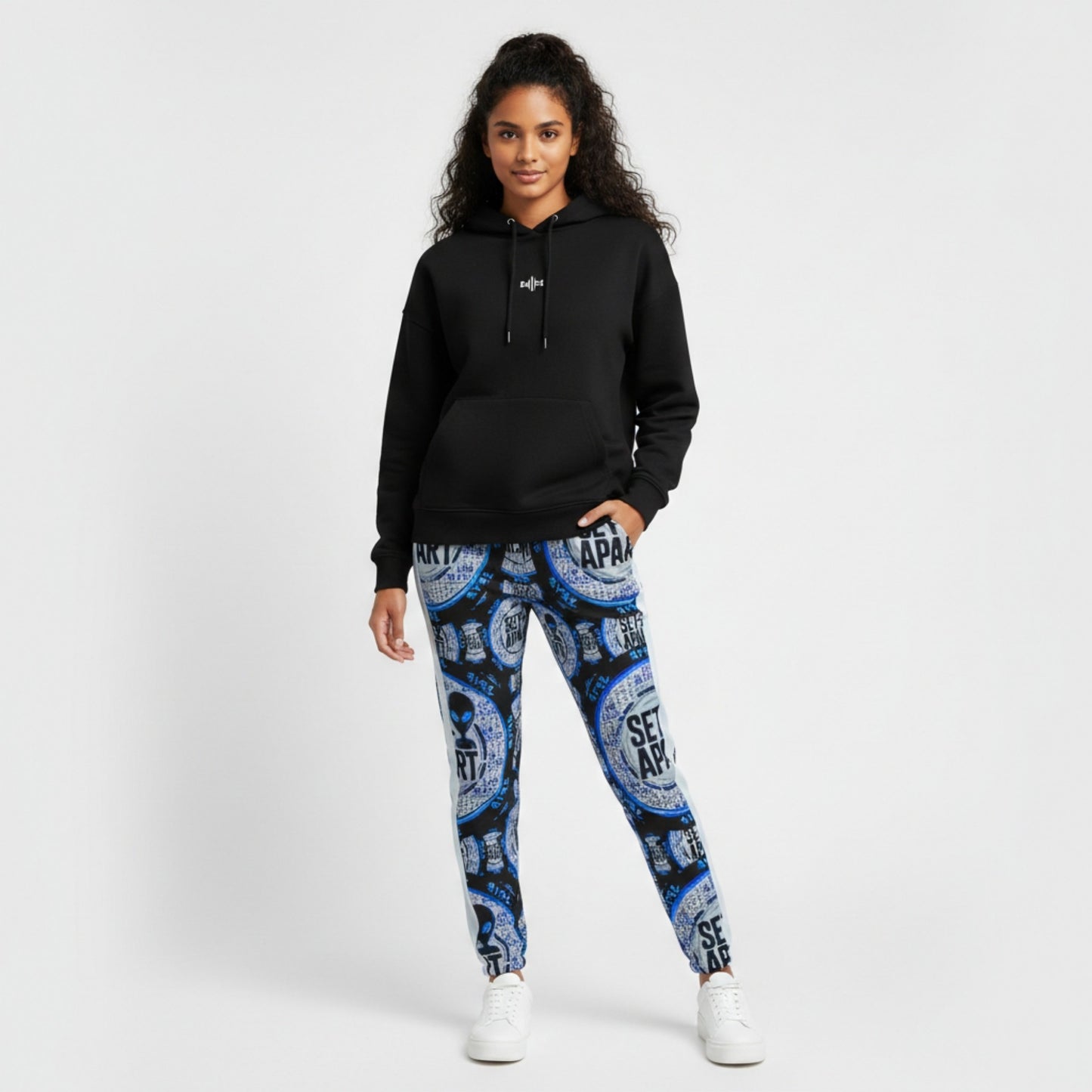 A woman in a black hoodie and blue graphic joggers with white sneakers. She is shown in front view against a white background.