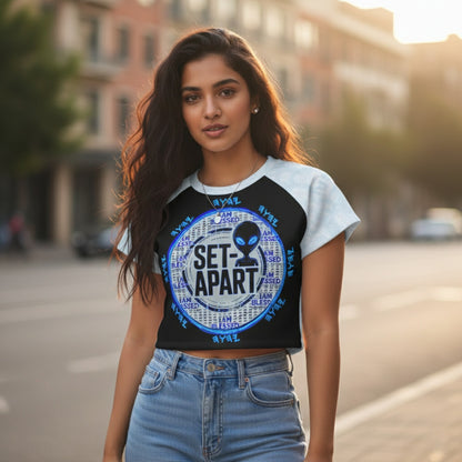 A woman standing on a city street at sunset wearing a "SET-APART" raglan-style crop top with a circular alien graphic and light-wash jeans.